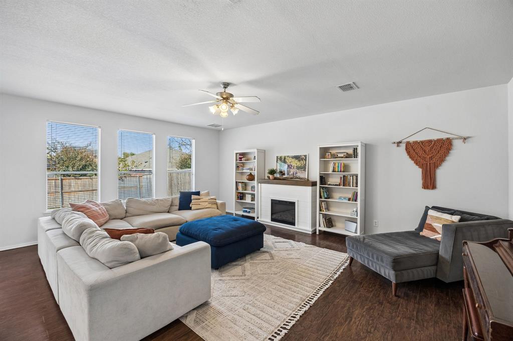 Spacious and welcoming, the living room features beautiful dark wood flooring, abundant natural light, and a soft, neutral color palette that creates an airy, comfortable atmosphere. Three large windows bring the outdoors in, while a charming fireplace framed by shelving creates an attractive focal point. The generous layout offers plenty of room for relaxing, gathering, and entertaining.