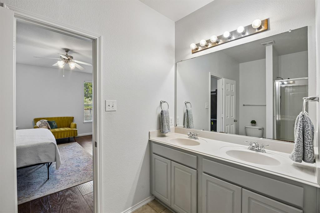 Designed with convenience in mind, this primary bathroom offers a long dual-sink vanity, generous counter space, and a large mirror illuminated by bright vanity lighting. Gray cabinetry adds a subtle contrast to the light countertops and neutral walls, while tile flooring provides a durable, easy-care surface. The layout offers functionality and comfort for daily routines.