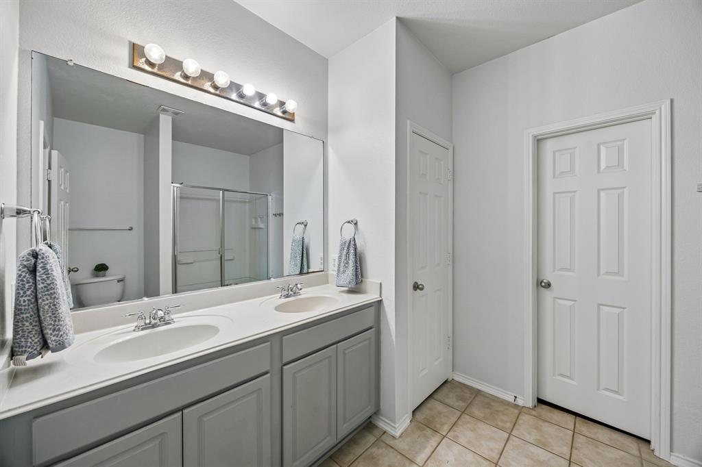 The primary bathroom features an extended dual-sink vanity with ample countertop space, providing added convenience for busy mornings and daily routines. Gray cabinetry, a large wall mirror, and bright vanity lighting contribute to a clean, functional design. Neutral tile flooring complements the surrounding finishes, while the adjoining shower area adds to the room's practicality.