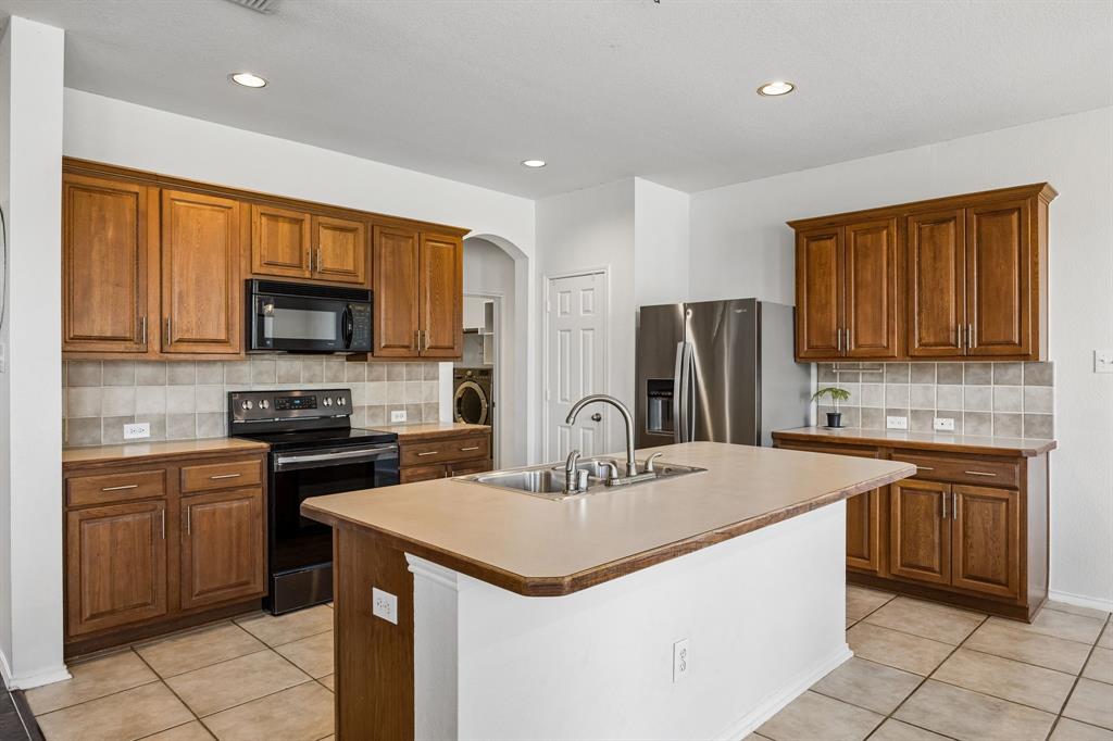 The kitchen offers generous cabinetry, ample countertop space, and a functional layout designed to support everyday cooking and entertaining. Warm wood cabinetry complements the neutral tile backsplash and flooring, creating a classic, inviting aesthetic. A spacious center island with an integrated sink provides additional workspace, while black and stainless steel appliances contribute a polished touch. Recessed lighting keeps the space bright, and the open connection to the adjoining dining and living areas enhances the home's easy flow.