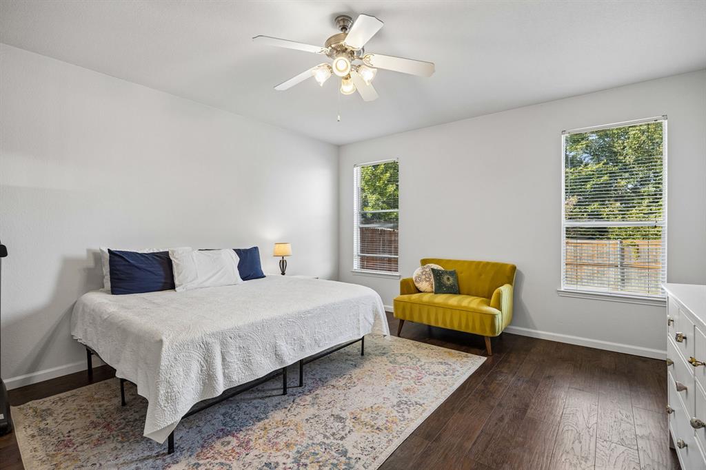 This spacious primary bedroom offers a comfortable retreat with neutral walls, warm wood-look flooring, and ample natural light streaming through two windows. A ceiling fan with integrated lighting adds everyday comfort, while the generous floor space allows for a variety of room arrangements. The light, neutral palette creates a welcoming atmosphere with plenty of potential to personalize the space.