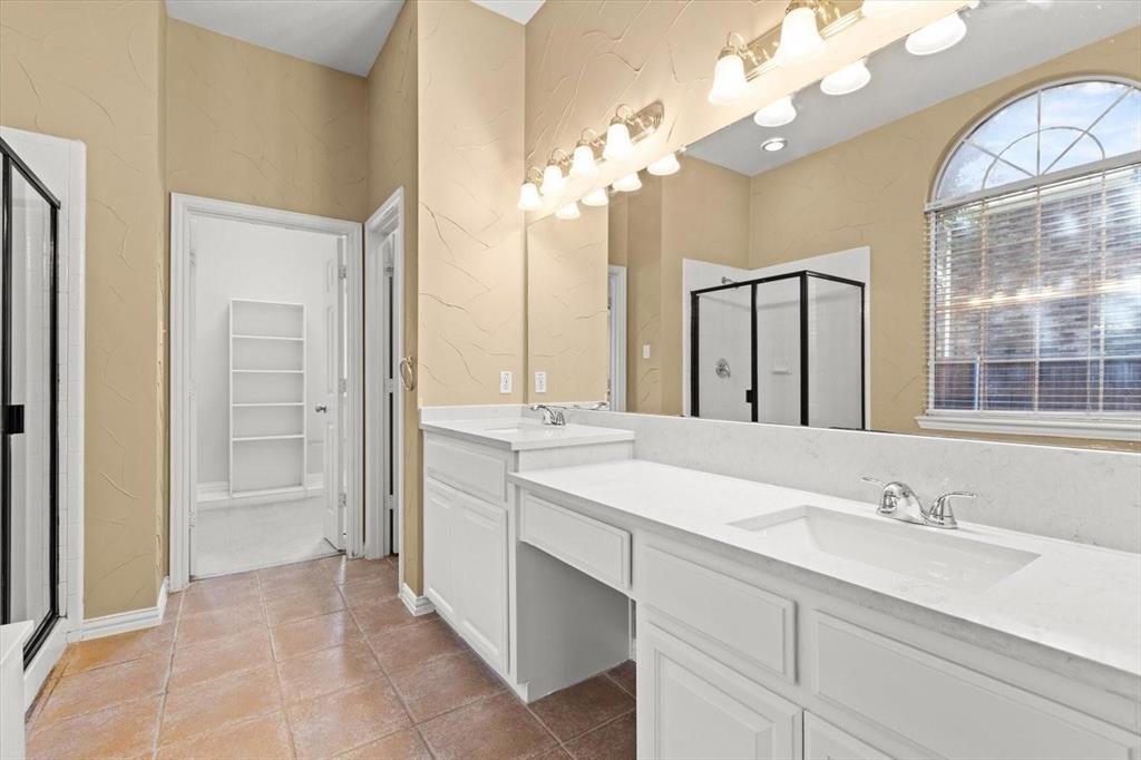 Bathroom featuring a dual-sink vanity with a solid-surface countertop, an arched window, tile flooring, and a glass-enclosed shower