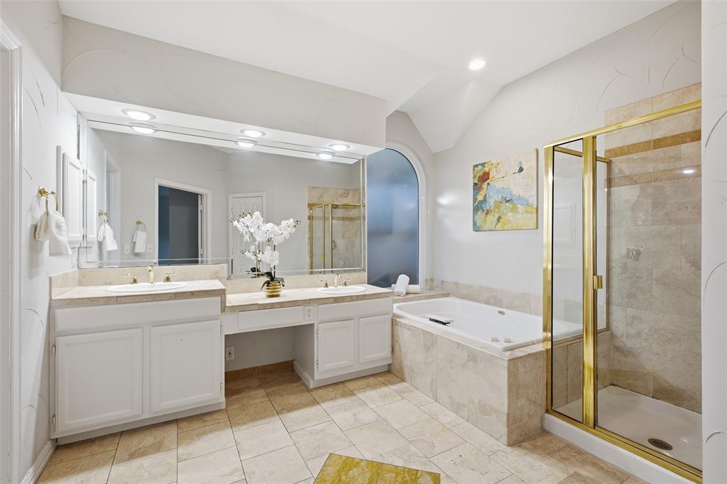 A spa-style primary bath with dual sinks, a built-in makeup vanity, a jetted soaking tub, a separate glass-enclosed shower and travertine-look finishes.