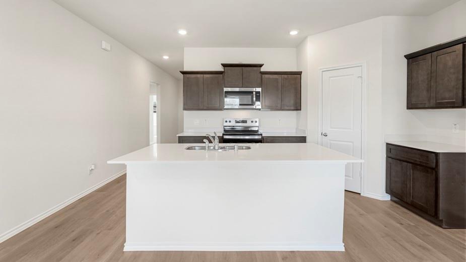 Kitchen featuring a large island with sink, stainless steel appliances, dark wood-finish cabinetry, light countertops, and wood-finish flooring