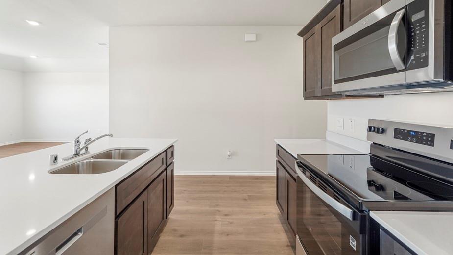 Kitchen featuring an island with a double basin sink, dark wood-finish cabinetry, light-toned countertops, a stainless steel microwave, and a black range