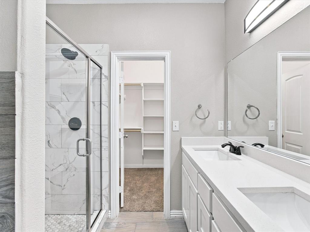Bathroom featuring a white double-sink vanity with a solid-surface countertop, walk-in shower with stone-tile surround and a frameless glass enclosure, and a built-in walk-in closet with integrated shelving