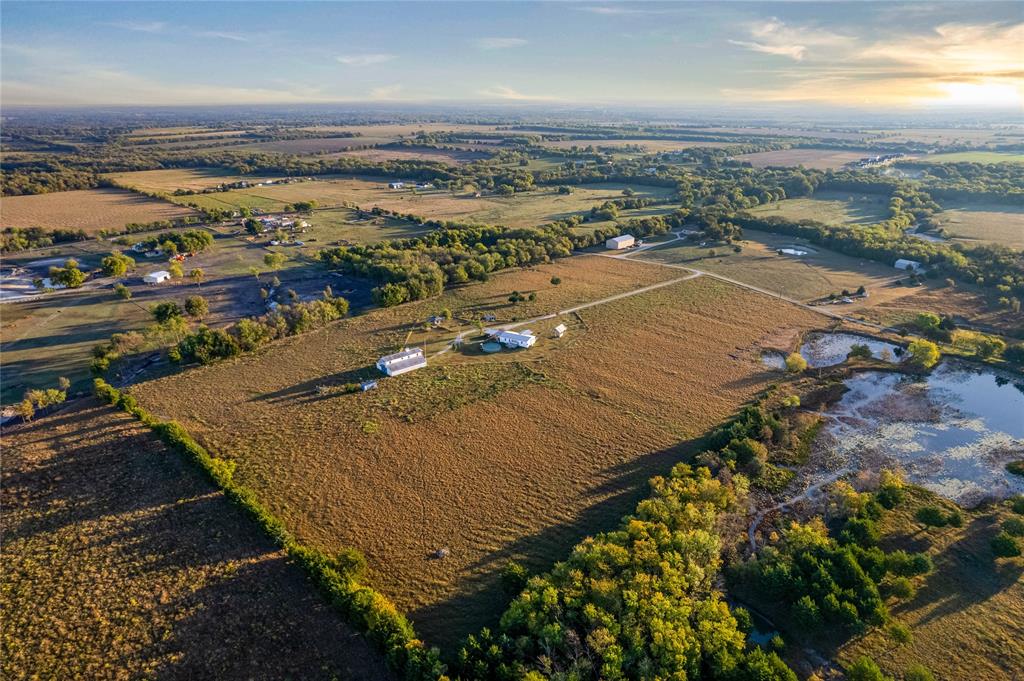 Aerial perspective of expansive acreage featuring open pasture, mature trees, a private pond, and multiple structural improvements