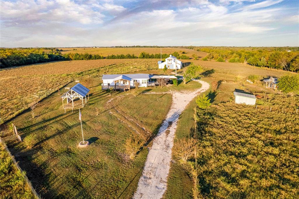 Aerial perspective of a multi-acre property featuring a single-story residence, a large metal-roofed barn, an open-sided pavilion, and a gravel driveway