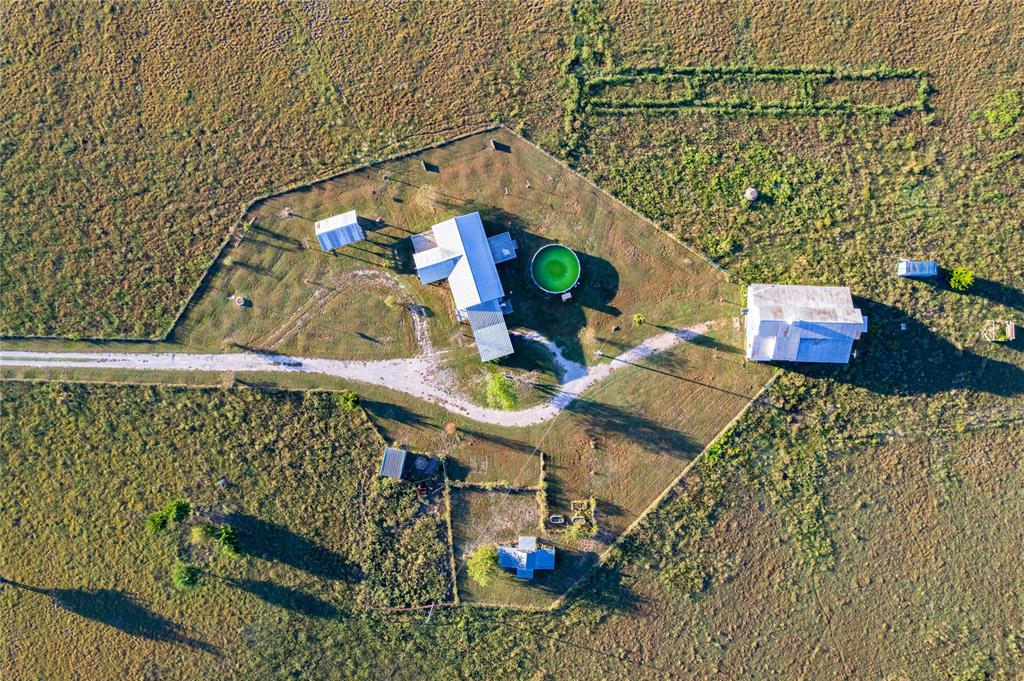 Aerial perspective of a multi-structure property featuring a main residence, additional outbuildings, a circular pool, and a gravel driveway, all enclosed within a fenced perimeter