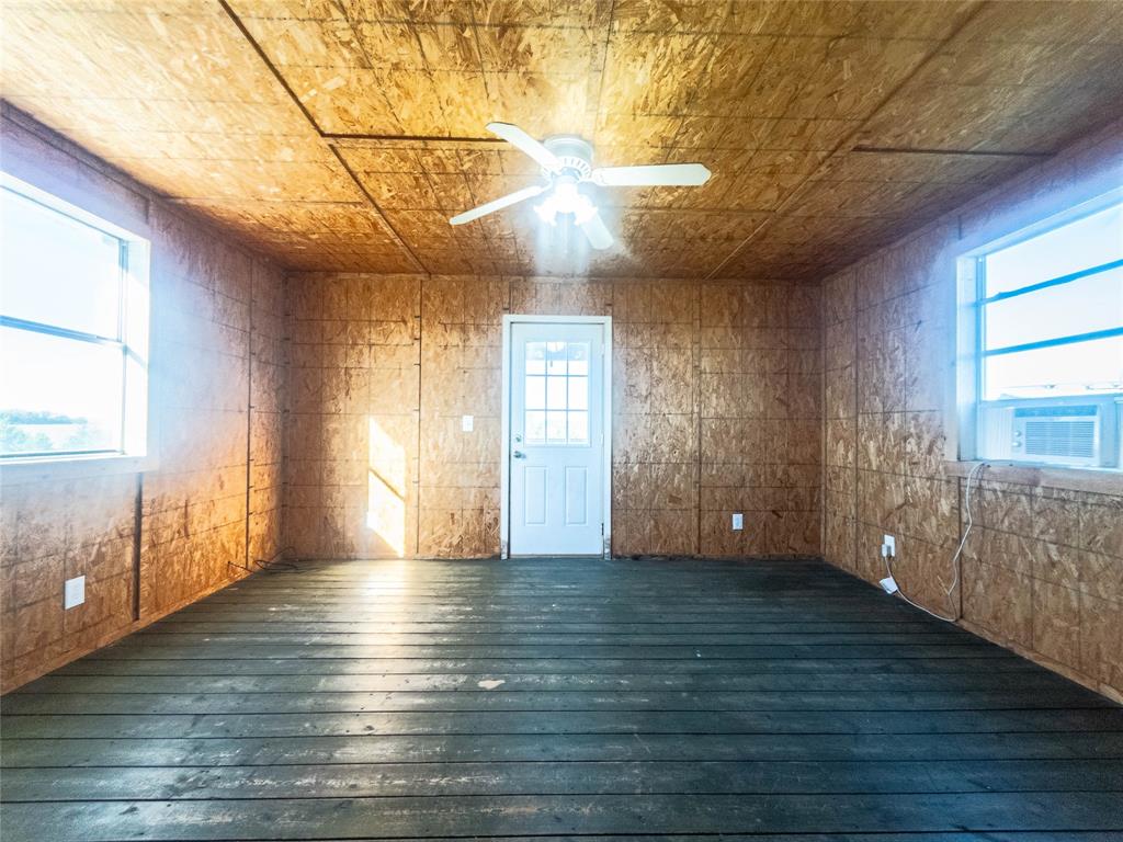 Interior room featuring oriented strand board wall and ceiling paneling, painted wood-plank flooring, dual windows, a central ceiling fan with integrated lighting, and a multi-pane exterior door