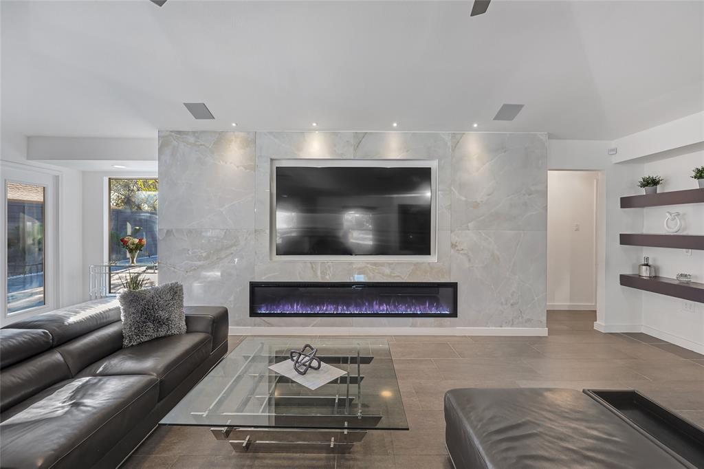 Large-format stone fireplace surround with a linear electric firebox, integrated wall-mounted television, recessed ceiling lighting, and built-in floating shelving