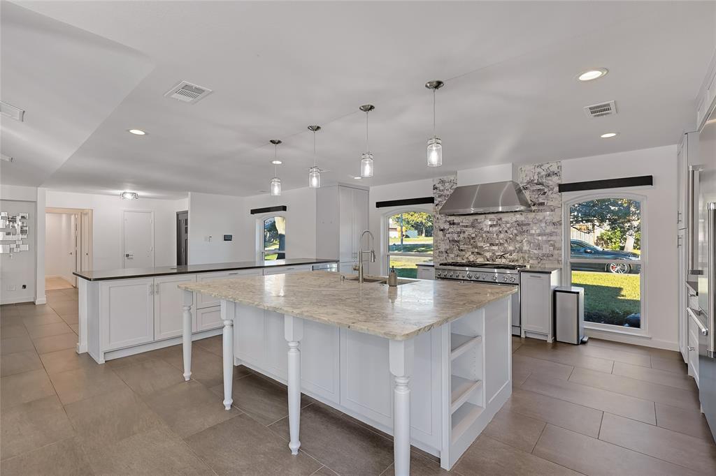 Kitchen featuring a large central island with stone countertop, stone-tiled backsplash, stainless steel range hood, pendant lighting, and oversized tile flooring