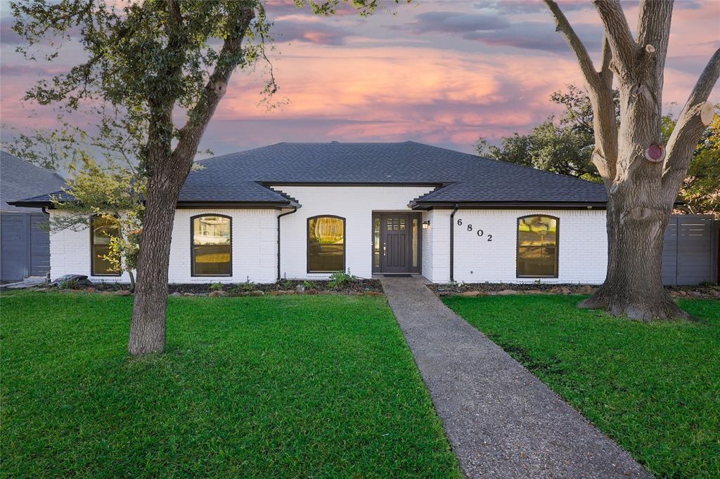 Single-story white brick exterior with a dark-tone shingle roof, arched windows, concrete walkway, and mature trees Single-story white brick exterior with a dark-tone shingle roof, arched windows, concrete walkway, and mature trees