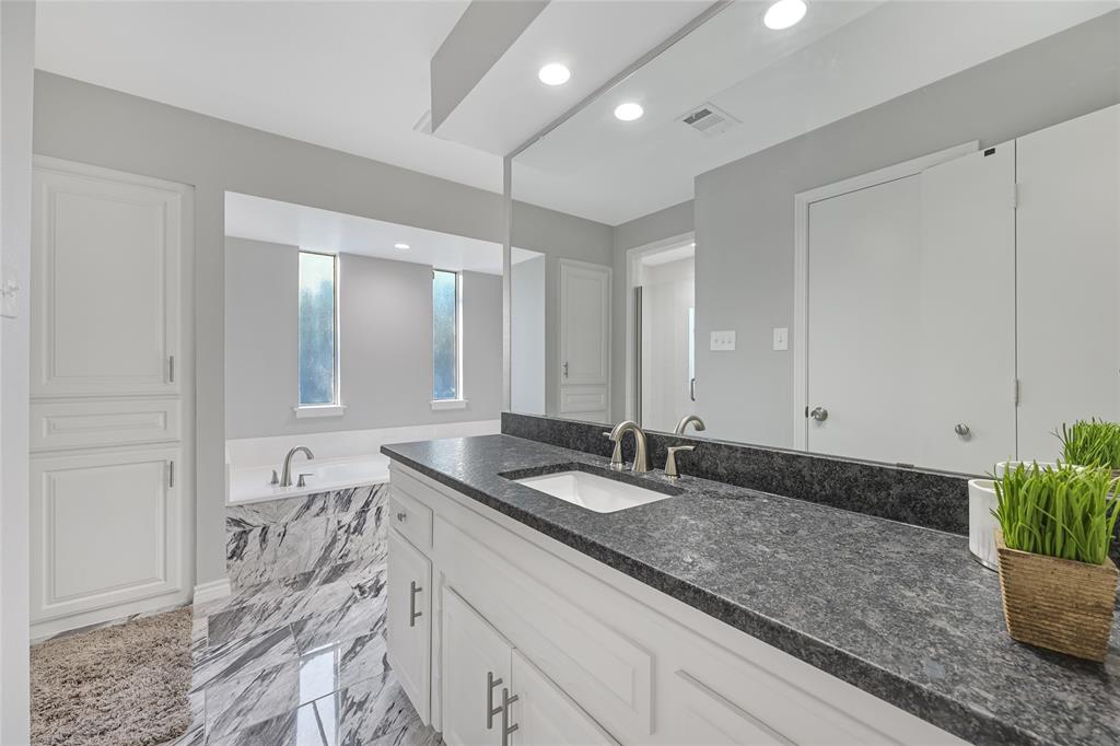 Bathroom featuring a white vanity with dark stone countertop, undermount rectangular sink, full-width mirror, and deep soaking tub surrounded by patterned stone tile