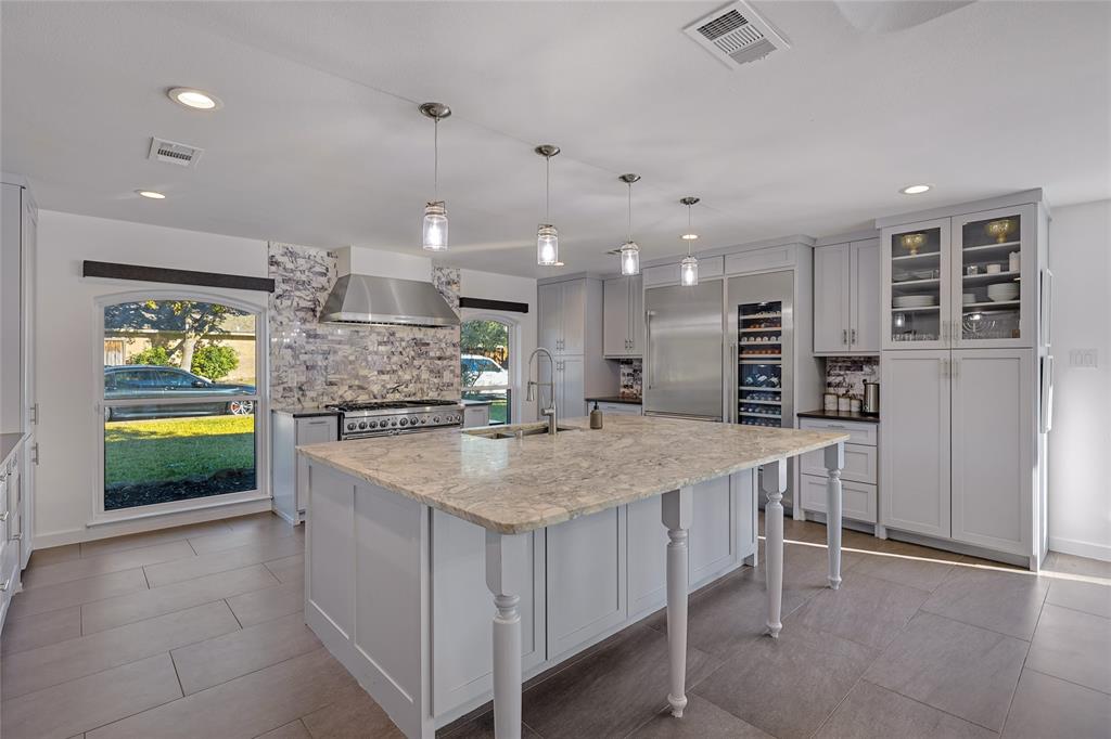 Kitchen featuring a central island with stone countertop, stainless steel professional-style range with matching vent hood, custom cabinetry with glass-front display sections, built-in wine refrigeration, and tile flooring