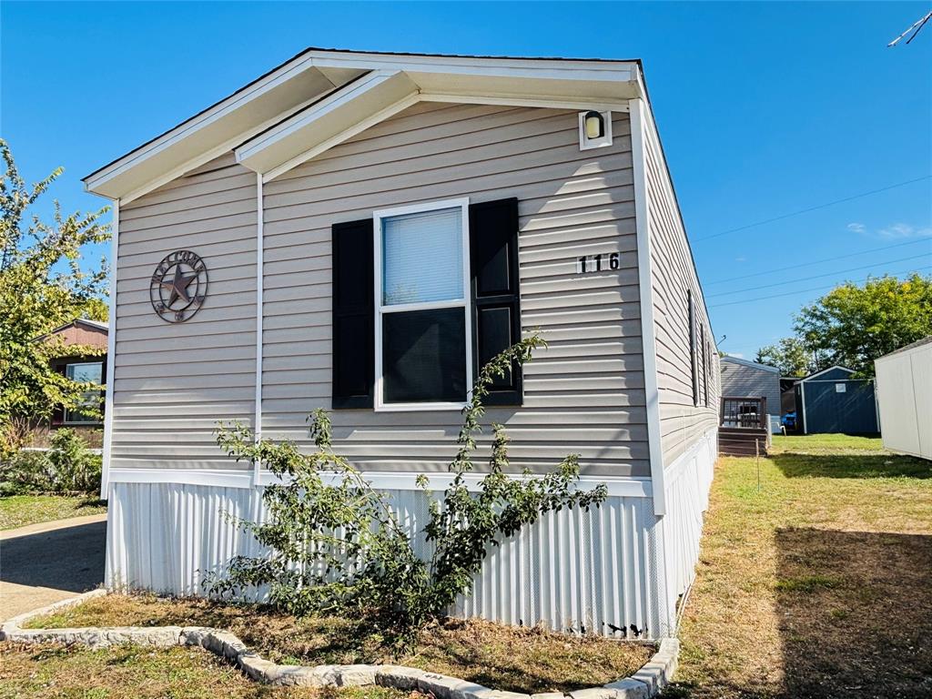 Exterior featuring light-tone horizontal vinyl siding, black window shutters, a white corrugated metal skirting, and a stone-bordered landscape edging