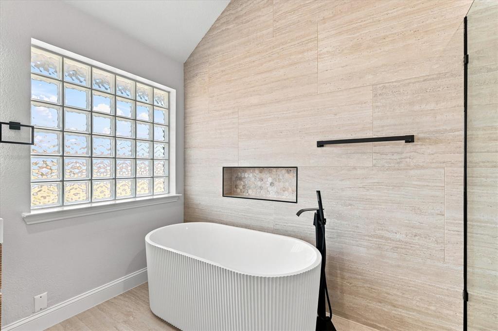 Freestanding fluted soaking tub with matte black floor-mounted faucet, floor-to-ceiling stone-finish tile wall, recessed niche, and glass block window