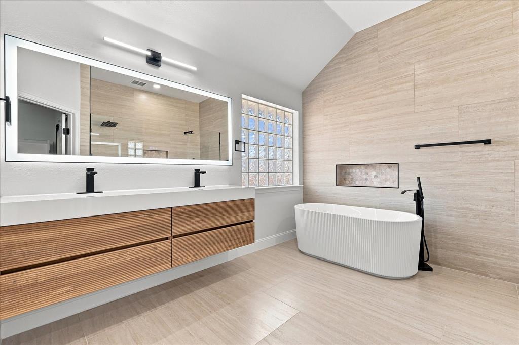 Floating double vanity with a dual-basin integrated countertop, ribbed freestanding soaking tub, matte black plumbing fixtures, large LED-backlit mirror, and full-height textured tile wall