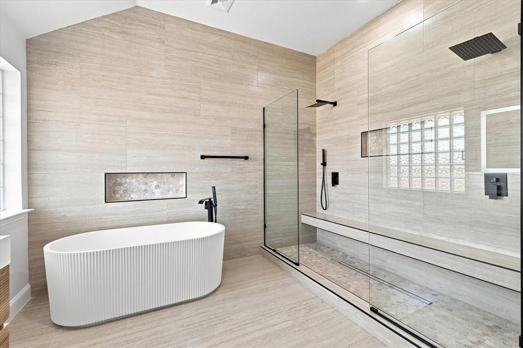 Freestanding fluted soaking tub, glass-enclosed walk-in shower with integrated bench seating, neutral linear tile walls, and matte black plumbing fixtures