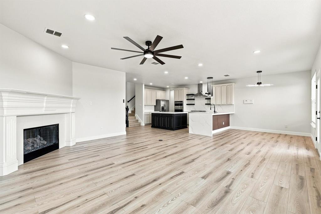 Open-concept interior featuring light wood-finish flooring, a white-mantel fireplace, a central kitchen island with a dark finish, integrated stainless steel appliances, and a multi-blade ceiling fan