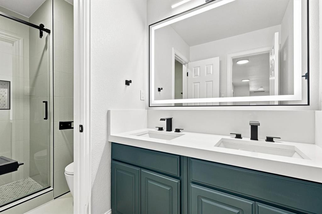 Dual-sink vanity with deep teal cabinetry and a solid-surface countertop, rectangular undermount sinks, matte black widespread faucets, and a large, integrated LED-backlit mirror