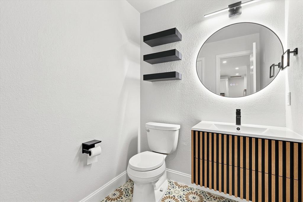Modern vanity with vertical wood-slat detailing, white rectangular basin, matte black faucet, illuminated circular wall mirror, and patterned tile flooring