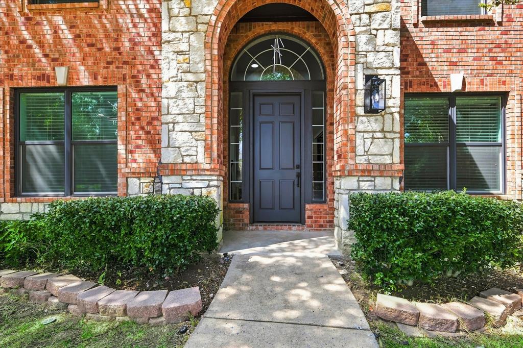 Exterior facade featuring a red brick and stone masonry veneer, arched entryway with a transom window, six-panel dark-finish entry door, and stone-bordered landscaping beds