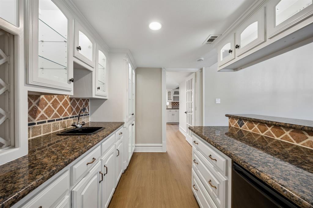 A closer look reveals the butler's panty distinctive craftsmanship, with beautifully detailed white cabinetry, glass-front display cabinets, and rich granite countertops. The decorative tile backsplash introduces an appealing blend of warm earth tones and geometric patterns, adding personality and visual interest. A convenient secondary sink and extensive counter space enhance functionality, while the thoughtful cabinet arrangement offers abundant storage and an attractive presentation.