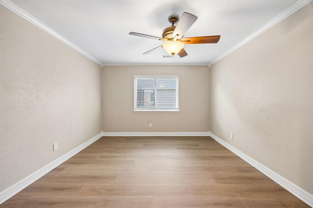 Enjoy the simple elegance of this inviting bedroom, featuring warm wood-look flooring, soft neutral walls, and decorative white crown molding. A window with white blinds brings in natural light, while the ceiling fan adds comfort and functionality. The elongated layout and generous wall space provide excellent versatility for a variety of uses.