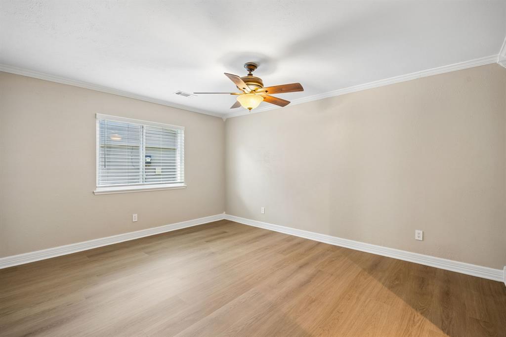 Warm wood-look flooring complements the soft neutral palette in this welcoming bedroom, while white baseboards and window trim add a polished finishing touch. Natural light filters through the window blinds, and the ceiling fan provides added comfort. The expansive wall space offers flexibility for a variety of bedroom layouts and design styles.