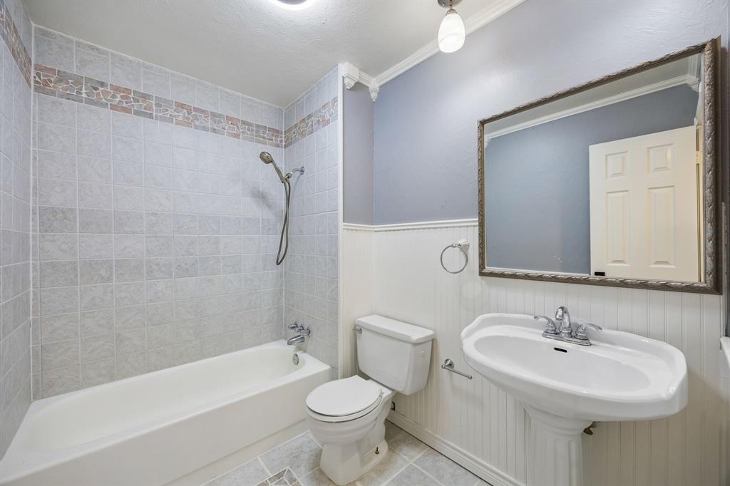 This well-appointed full bathroom features a classic white bathtub and shower combination surrounded by light-colored tile, complemented by a decorative accent border for added visual appeal. White beadboard wainscoting, a pedestal-style sink, and a generously sized mirror create a timeless aesthetic, while the soft blue-gray walls and coordinating tile flooring contribute to a fresh, inviting atmosphere.