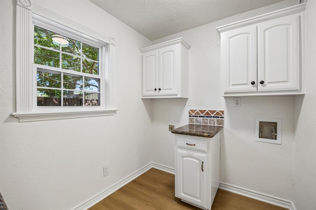 A charming and thoughtfully arranged utility room features white cabinetry, granite countertops, and coordinating tile accents that carry the home's distinctive design throughout. A large window welcomes natural light and offers a pleasant view of the outdoors, while the dedicated washer and dryer connections provide a convenient space for everyday laundry needs. Warm wood-look flooring and classic trim complete this bright, functional area.
