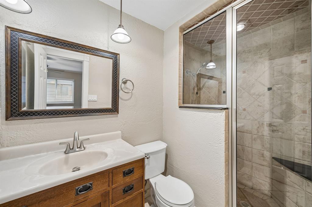 A well-appointed bathroom features a warm wood-tone vanity with ample drawer storage, a light countertop, and coordinating fixtures. The expansive framed mirror enhances the sense of space, while the tiled walk-in shower showcases a decorative tile pattern, glass enclosure, and built-in bench. Neutral finishes and thoughtful details create a comfortable, functional space.