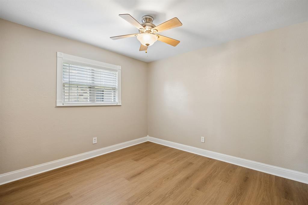 A bright and inviting bedroom showcases warm wood-look flooring, soft neutral walls, crisp white trim, and a charming ceiling fan with integrated lighting. A well-positioned window with white blinds welcomes natural light, creating a comfortable and versatile space ready to suit a variety of needs.