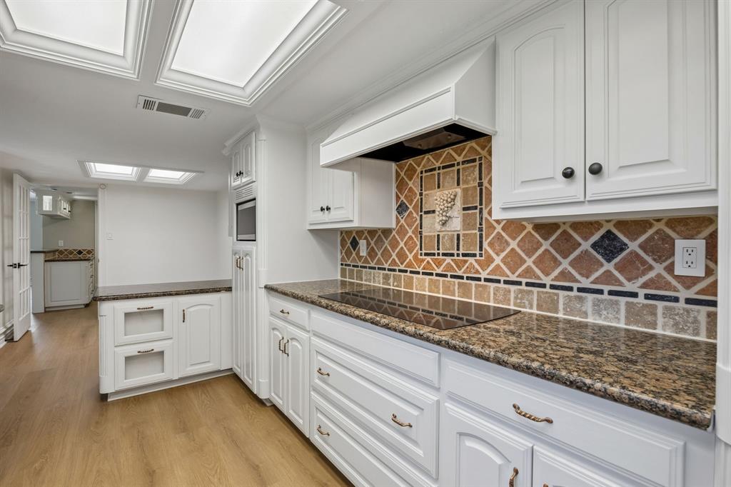 Another view of the beautifully appointed kitchen showcasing crisp white cabinetry with decorative molding, rich granite countertops, and a distinctive multicolored tile backsplash that adds warmth and character. A sleek cooktop, built-in microwave, and custom range hood create a functional cooking space, while recessed ceiling panels and abundant cabinetry enhance the room's timeless appeal. Warm wood-look flooring flows throughout, complementing the inviting neutral palette.