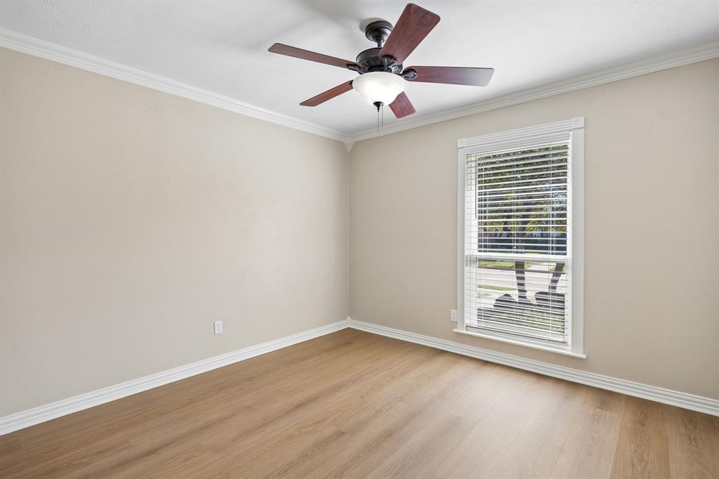 A comfortable bedroom offers a peaceful retreat with soft neutral walls, warm wood-look flooring, crisp white baseboards, and elegant crown molding. A ceiling fan with integrated lighting adds everyday comfort, while the generously sized window welcomes natural light and provides views of the surrounding greenery. The clean lines and versatile color palette create an inviting canvas for a variety of design styles.