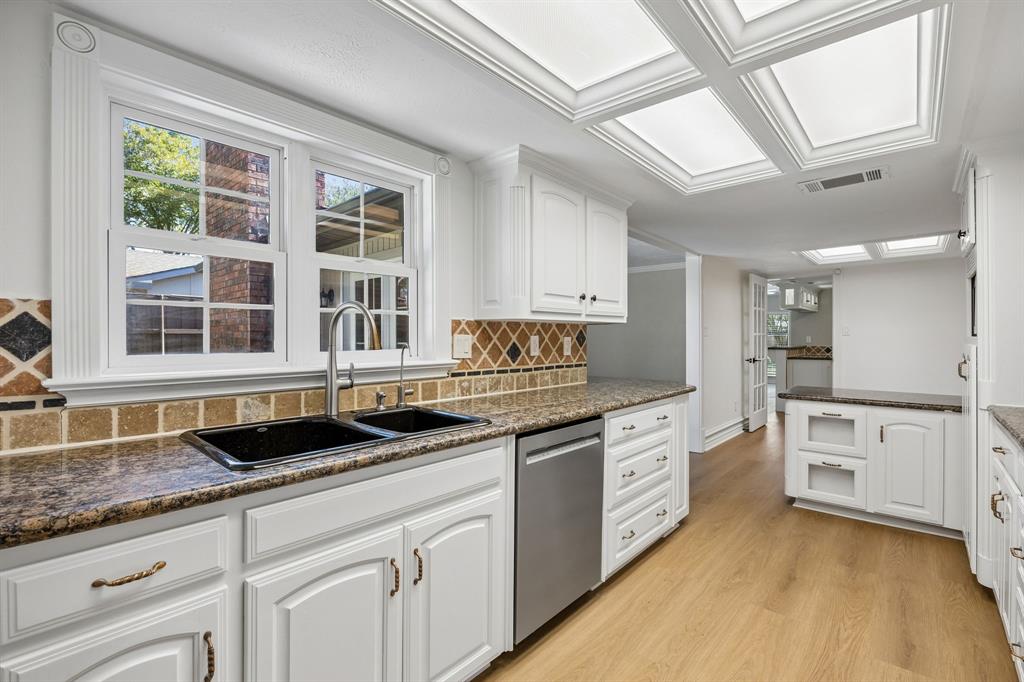 Natural light pours through the kitchen window, illuminating the expansive countertops, deep double-basin sink, and classic white cabinetry. Decorative tile backsplash details add texture and visual interest, while the stainless steel dishwasher and ample drawer storage enhance everyday practicality. The open sightlines and warm flooring create a welcoming kitchen that balances traditional character with functional design.