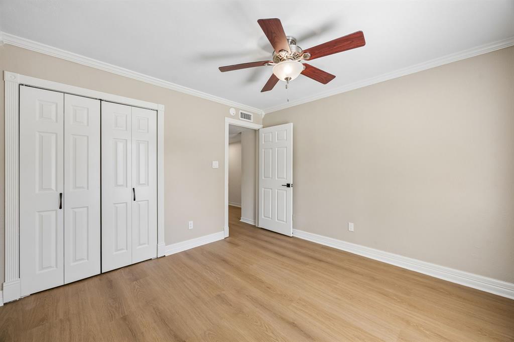 A spacious bedroom showcases warm wood-look flooring, soft neutral walls, and classic white architectural trim that lends a polished, cohesive finish. A ceiling fan with integrated lighting adds comfort, while the wide closet with traditional paneled doors offers practical storage. The generous open floor area and understated color scheme create a flexible space ready to accommodate a variety of uses and design preferences.
