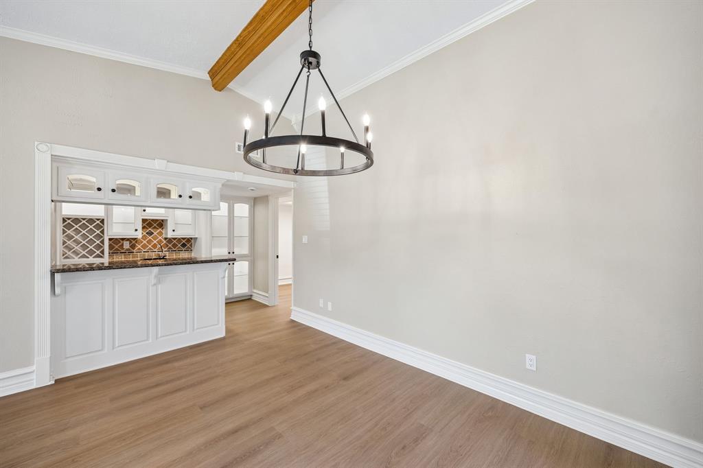 Architectural details take center stage in this inviting dining space, where a statement chandelier hangs beneath a warm wood beam. The built-in serving bar features classic white cabinetry, dark countertops, and decorative tile accents, creating a distinctive focal point. An open connection to the kitchen enhances the home's natural flow, making entertaining effortless.