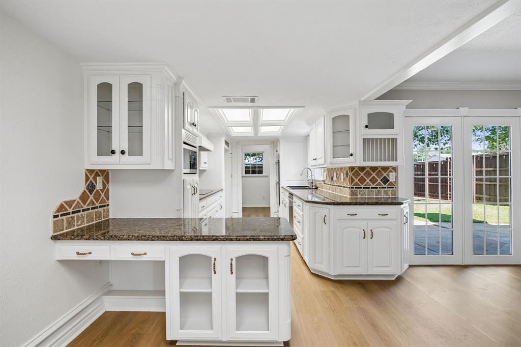 A bright, thoughtfully arranged kitchen features abundant white cabinetry, richly patterned countertops, and distinctive diamond-pattern tile backsplash detailing. A central peninsula provides additional workspace and storage, while glass-front cabinet doors and decorative millwork add character. Overhead natural-light panels brighten the space, and French doors offer convenient access to the backyard.