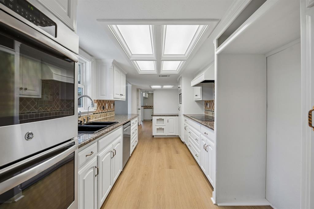 Designed with functionality and classic appeal in mind, the kitchen offers extensive cabinetry, generous counter space, and an efficient galley-style layout. White cabinetry creates a crisp, timeless backdrop for the contrasting countertops and decorative tile backsplash, while stainless steel appliances add a contemporary touch. Light wood-look flooring carries warmth throughout the space, and the abundant storage provides excellent everyday convenience.
