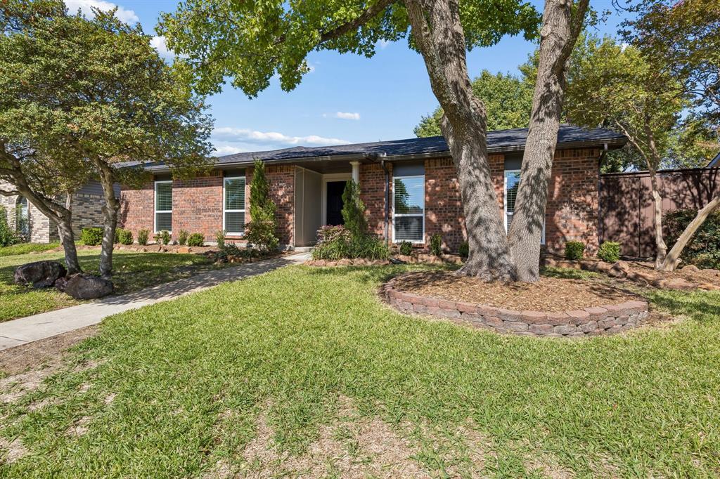 Mature trees provide a picturesque canopy over the front lawn, complementing the home's warm brick facade and established landscaping. A graceful walkway leads to the covered entrance, while a raised stone garden bed adds character and dimension to the beautifully shaded front yard.