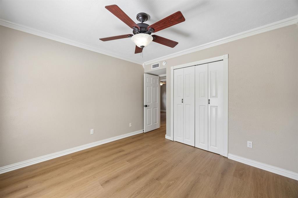 This inviting bedroom combines understated elegance with practical features, including warm wood-look flooring, decorative crown molding, and a ceiling fan with integrated lighting. A spacious closet with classic white folding doors provides convenient storage, while the neutral finishes and open floor area allow flexibility for furniture placement. The bright, uncluttered presentation highlights the room's comfortable proportions and timeless appeal.