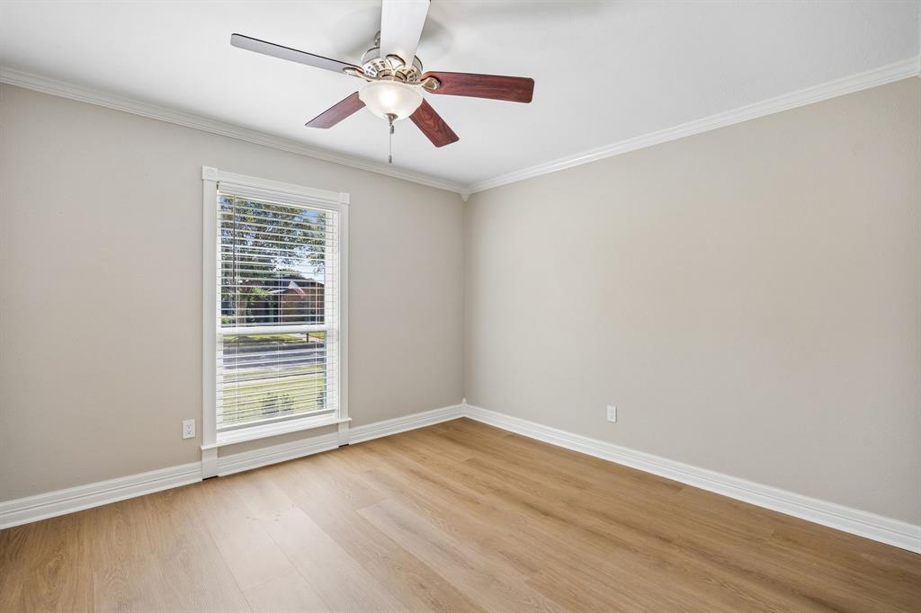 Bright and welcoming, this bedroom features a soothing neutral palette, warm wood-look flooring, crisp white trim, and an attractive ceiling fan with integrated lighting. A large window framed by decorative molding allows natural light to fill the space while offering views of the outdoors. The simple, elegant finishes create a versatile retreat suited to a variety of personal styles.
