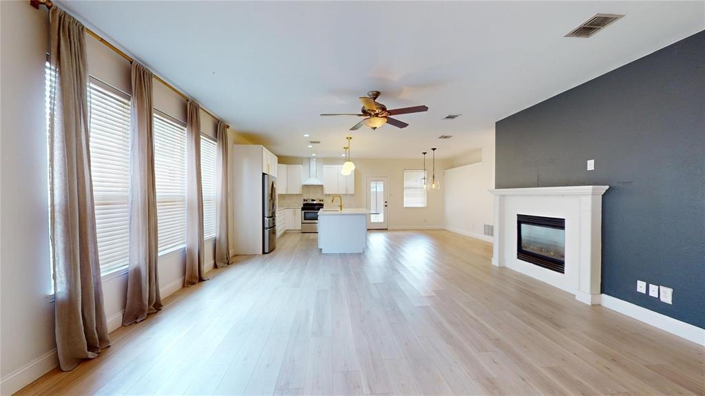 Open-concept living space featuring wood-finish flooring, a wall-mounted electric fireplace with a white mantel, kitchen island with quartz-style countertop, white cabinetry, and large windows with natural light
