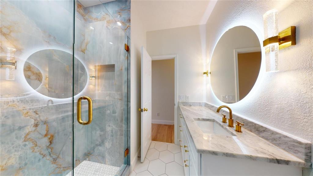 Bathroom featuring a walk-in shower with large-format decorative wall tile, brass-finish hardware, a stone countertop vanity with integrated sink, a backlit circular mirror, and hexagonal floor tile