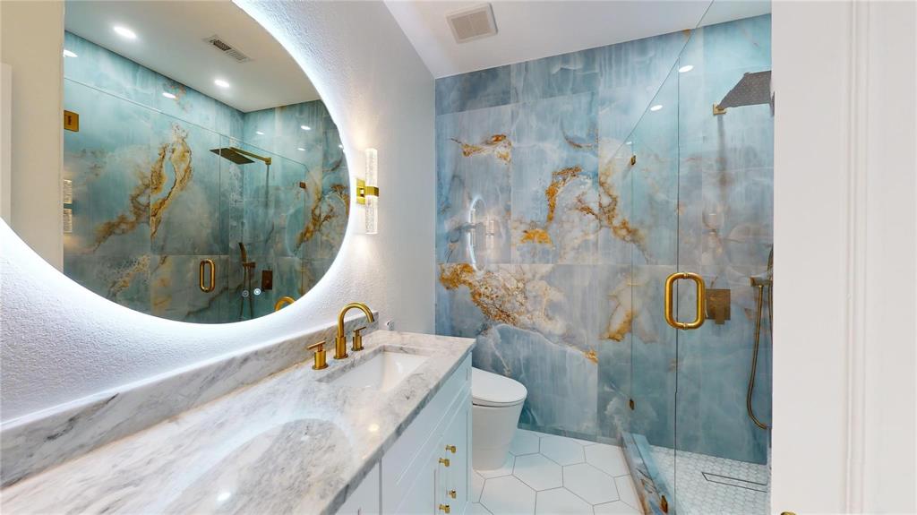 Bathroom featuring large-format blue and gold-toned wall tile, a frameless glass-enclosed shower, a backlit circular mirror, brass-finish hardware, and a stone-surface vanity
