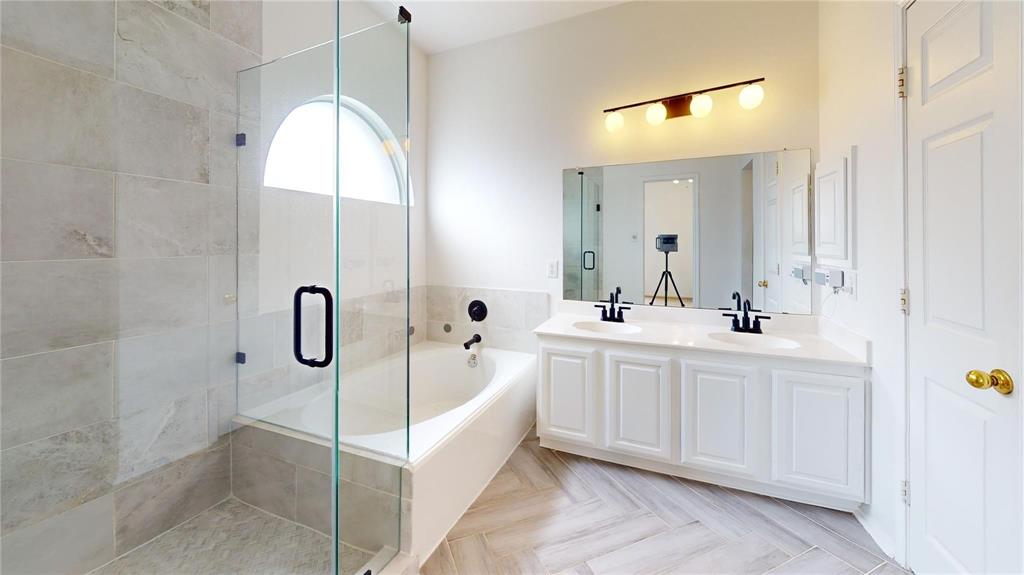Dual-sink vanity with white cabinetry, glass-enclosed shower with tile surround, soaking tub, arched window, and herringbone-patterned tile flooring