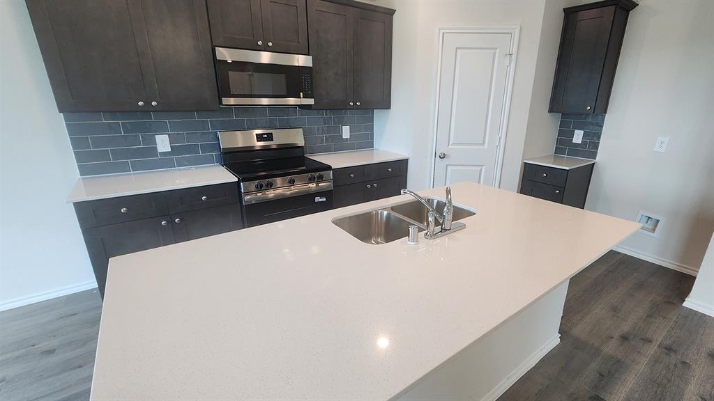 Dark wood-finish cabinetry, stainless steel range and microwave, light-toned countertops, subway tile backsplash, and a kitchen island with an undermount double-basin sink