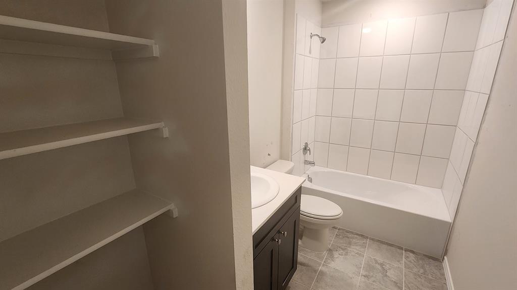 Built-in shelving, dark wood-finish vanity with countertop, undermount sink, white tile shower surround, and gray-toned floor tiling